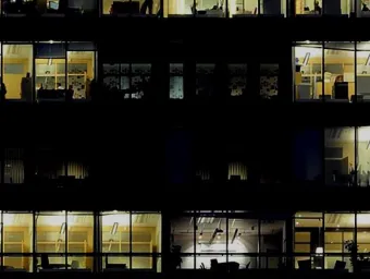 It is a dark cityscape, with the darkness only punctured by small pools of light from inside an empty office, creating a sense of foreboding